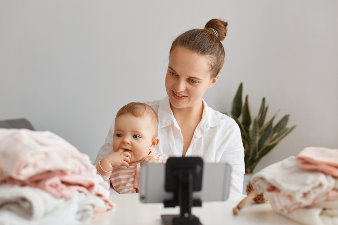 Beautiful Young Adult Woman Blogger Sitting at Table with Toddler Kid and Recording Video for Her Vlog, Using Mobile Phone on Tripod, Making Content with Her Daughter.